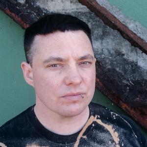 Headshot of Malic Amalya in front of a grey-green wall and a rusted, diagonal, support beam.