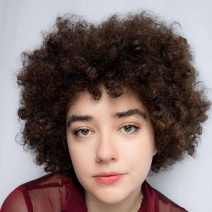 A young person with medium length brown coily hair looks intently into the camera, face relaxed