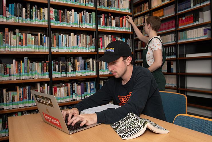 A student works on a laptop in the library while another selects a book