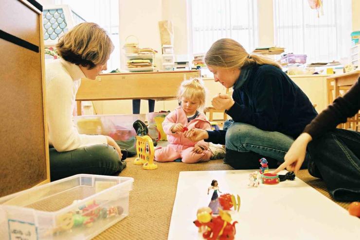 Two adults sitting on the floor of a colorful playroom surrounded by toys, engaging a young child