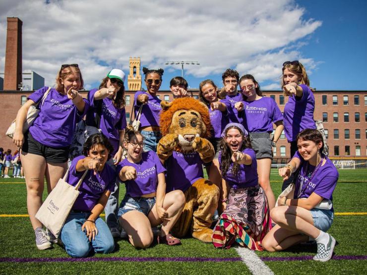 Eleven students in matching Emerson College shirts excitedly pose with the Emerson lion mascot