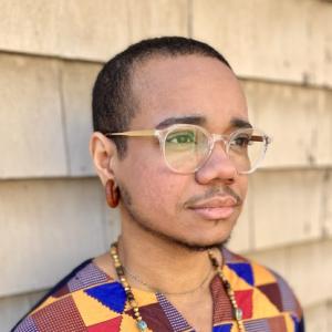 Headshot of Eddie Maisonet, a Black nonbinary person with plastic frame glasses, looks off at an angle with a neutral expression.