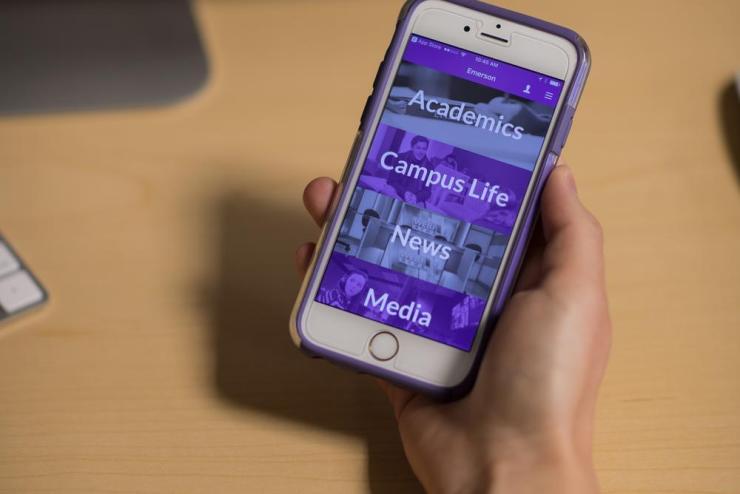 A hand holding an iPhone displaying the Emerson College App